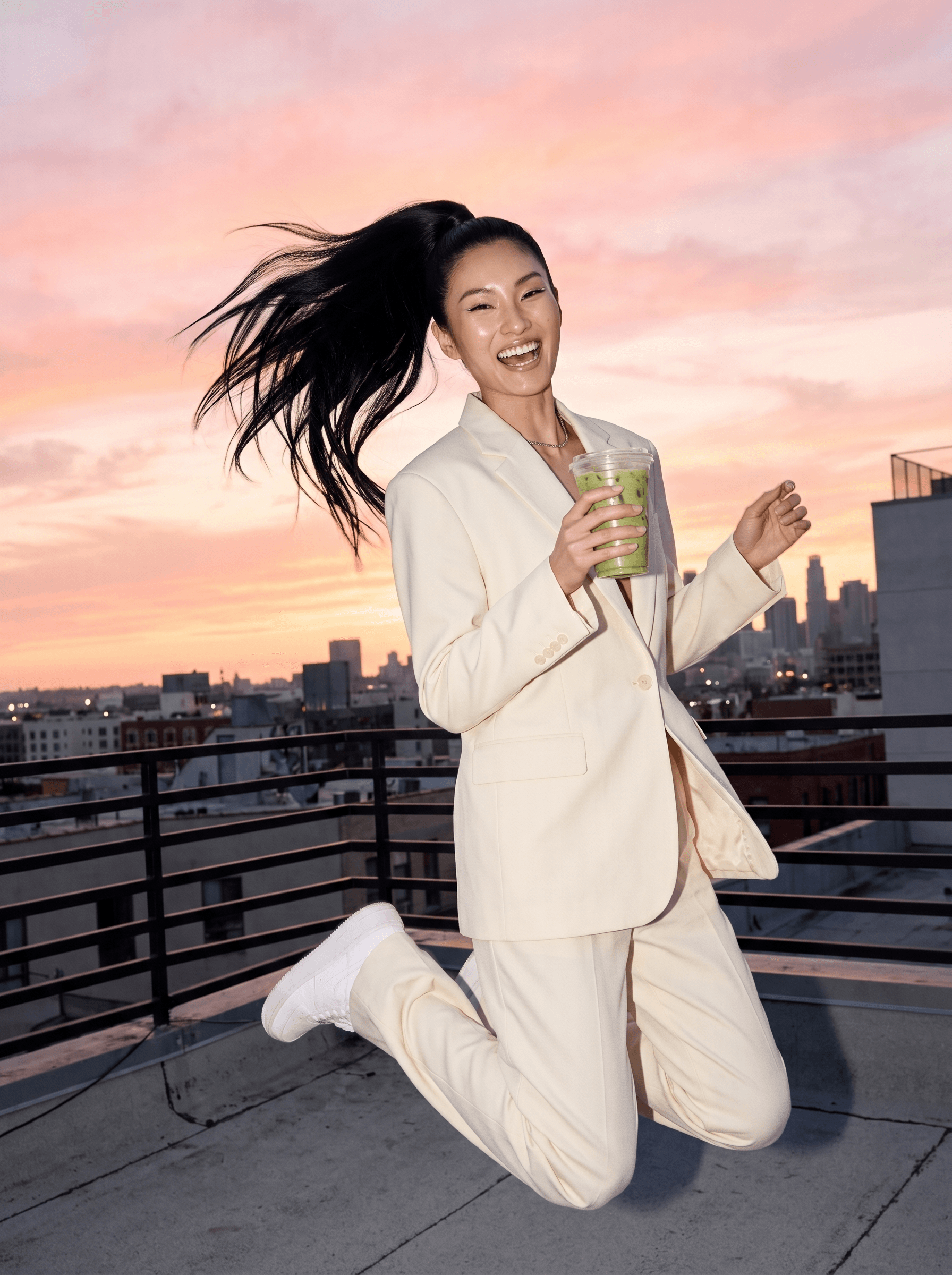 Woman with a long black ponytail jumping on a rooftop with iced matcha